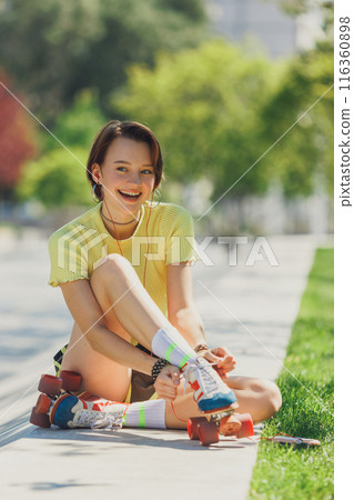 Energetic young lady in yellow crop top, seated on concrete ramp, adjusting her earphones with joyful expression. Energetic young lady in yellow crop top, seated on concrete ramp, adjusting her earphones with joyful expression. 116360898