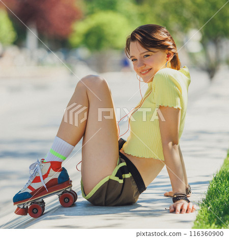 Happy woman in yellow top and brown shorts sitting on park ramp, enjoying outdoors with her retro roller skates and earphones. 116360900
