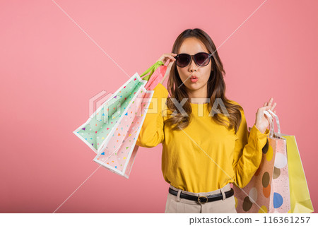 In a studio shot on pink background, a joyful Asian woman, elegantly holding shopping bags, radiates happiness. She's the portrait of a satisfied shopper. black friday 116361257
