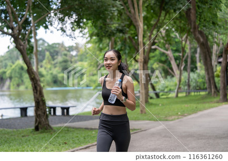 Young Woman Jogging in Park with Water Bottle, Enjoying Outdoors Exercise in Green Natural Environment 116361260