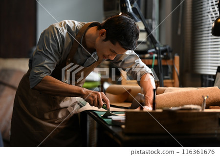 Small business craftsman in apron carefully cutting piece of leather in his workshop Small business craftsman in apron carefully cutting piece of leather in his workshop 116361676