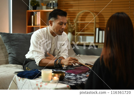 Fortune teller during a reading session talking to client at table surrounded by candles and various spiritual items Fortune teller during a reading session talking to client at table surrounded by candles and various spiritual items 116361720