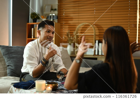 Fortune teller during a reading session talking to client at table surrounded by candles and various spiritual items Fortune teller during a reading session talking to client at table surrounded by candles and various spiritual items 116361726