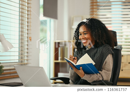 Smiling African American woman wearing headphones using laptop at home and writing notes Smiling African American woman wearing headphones using laptop at home and writing notes 116361737