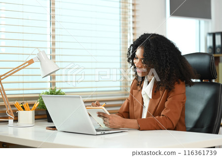 Smiling African American businesswoman in brown jacket working with laptop and making note at office desk Smiling African American businesswoman in brown jacket working with laptop and making note at office desk 116361739