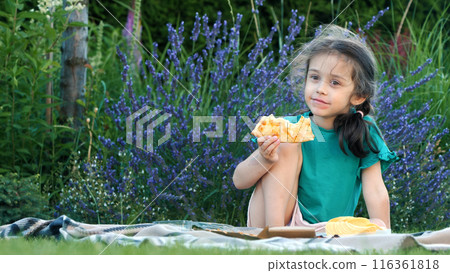 A happy girl eagerly eating a slice of pizza while sitting on a blanket at an impromptu picnic. 116361818