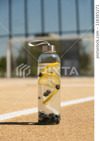 Transparent glass bottle of water or isotonic with lemon and berry on outdoor soccer sports ground. Concept water balance for workout. Transparent glass bottle of water or isotonic with lemon and berry on outdoor soccer sports ground. Concept water balance for workout. 116365271