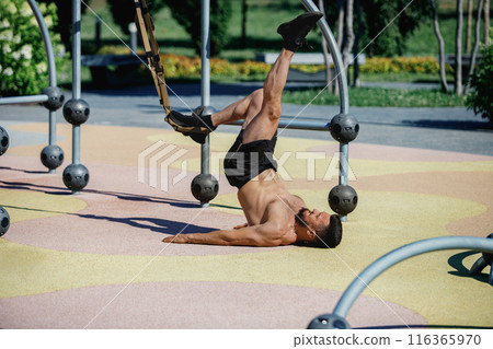 Strong shirtless man lying on ground in park and training with trx straps, doing exercises. Outdoor intense training 116365970