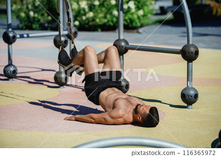 Athletic man in black shorts exercising with trx straps, lying on the ground, lifting his legs, park training on sunny day. 116365972