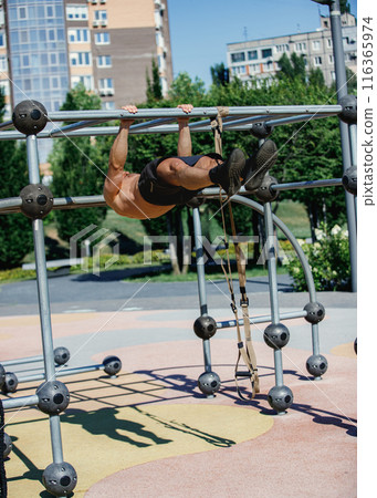 Athletic man in black shorts doing pull-ups on metal frame in park, showcasing muscular body. Early morning training 116365974