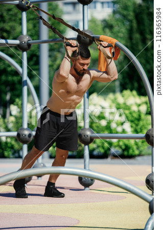 Shirtless man in black shorts working out with suspension straps, doping intense trx exercises in outdoor park, under sunny weather. Shirtless man in black shorts working out with suspension straps, doping intense trx exercises in outdoor park, under sunny weather. 116365984