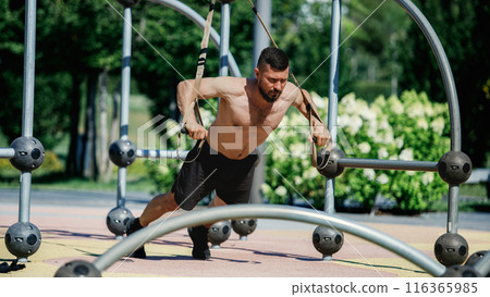 Athletic man in park, training shirtless, exercising with suspension straps attached to metal frame. TRX training session. Push ups Athletic man in park, training shirtless, exercising with suspension straps attached to metal frame. TRX training session. Push ups 116365985