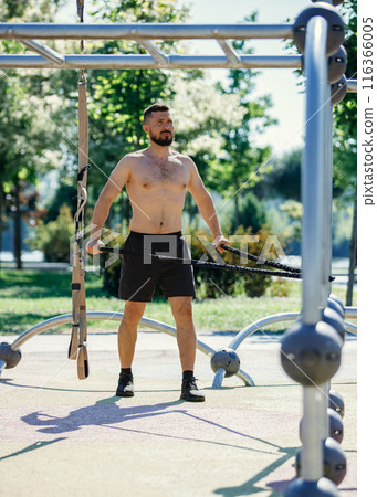 Shirtless man exercising with resistance bands in outdoor park, wearing black shorts, focusing on his workout. Muscular body shape Shirtless man exercising with resistance bands in outdoor park, wearing black shorts, focusing on his workout. Muscular body shape 116366005