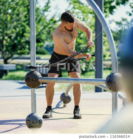 Bearded man performing resistance band exercises shirtless, in black shorts, at outdoor fitness park on sunny day. Bearded man performing resistance band exercises shirtless, in black shorts, at outdoor fitness park on sunny day. 116366014