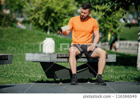 Athletic man wearing orange and black sportswear sitting on bench, using shaker bottle for making proteins shaker. Outdoor training Athletic man wearing orange and black sportswear sitting on bench, using shaker bottle for making proteins shaker. Outdoor training 116366049