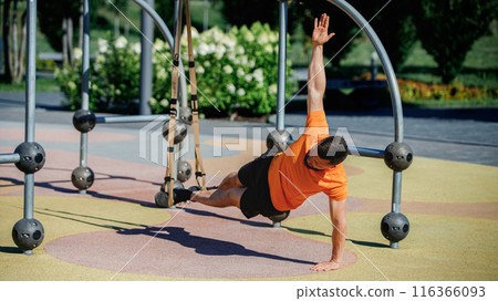 Muscular man performing TRX side plank outdoors, balancing on one arm with his feet suspended. Outdoor intense training Muscular man performing TRX side plank outdoors, balancing on one arm with his feet suspended. Outdoor intense training 116366093