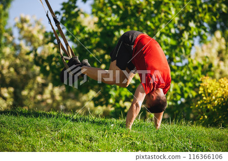 Fit athlete, muscular man in sportswear training outdoors in park, performing an intense TRX pike workout on a grassy ground Fit athlete, muscular man in sportswear training outdoors in park, performing an intense TRX pike workout on a grassy ground 116366106
