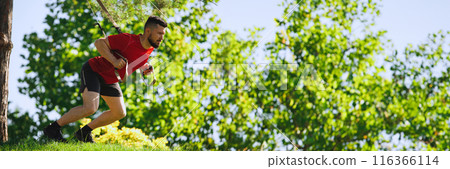 Fit athlete, man in red t-shirt performing TRX exercises outdoors with straps. Muscular man doing squats exercises in greenery park 116366114