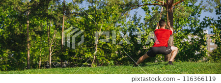 Back view. Athletic man in sportswear training outdoors, doing trx workout in greenery park, doing squats exercises. Back view. Athletic man in sportswear training outdoors, doing trx workout in greenery park, doing squats exercises. 116366119