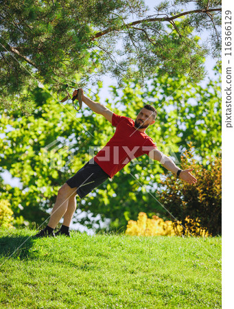 TRX workout session. Muscular bearded man in red t-shirt training outdoors in greenery park, using trx straps for exercises. TRX workout session. Muscular bearded man in red t-shirt training outdoors in greenery park, using trx straps for exercises. 116366129