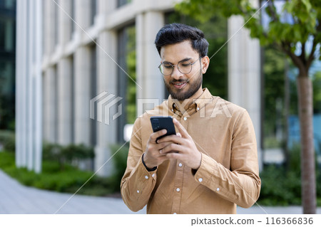 Young businessman using smartphone outside office building, focused on device. Casual attire, modern corporate environment, professional lifestyle. Urban exterior, connected 116366836