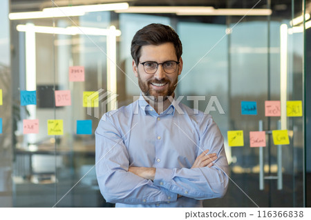 A man with glasses and a beard is smiling in front of a wall covered in sticky notes. The notes are yellow and pink, and they seem to be related to a project or a task 116366838