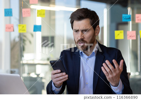 Sad disappointed businessman reading news on phone, man in business suit using app on smartphone, looking upset, received bad news online in email, working with laptop inside office. 116366890