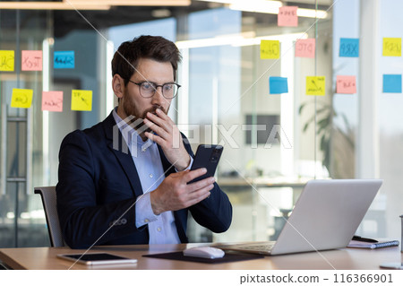 Sad disappointed businessman reading news on phone, man in business suit using app on smartphone, looking upset, received bad news online in email, working with laptop inside office. Sad disappointed businessman reading news on phone, man in business suit using app on smartphone, looking upset, received bad news online in email, working with laptop inside office. 116366901