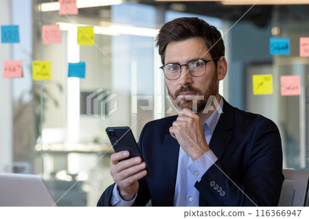 Mature businessman in a modern office setting using a smartphone. He appears thoughtful, wearing glasses and a dark suit, surrounded by colorful sticky notes. Mature businessman in a modern office setting using a smartphone. He appears thoughtful, wearing glasses and a dark suit, surrounded by colorful sticky notes. 116366947