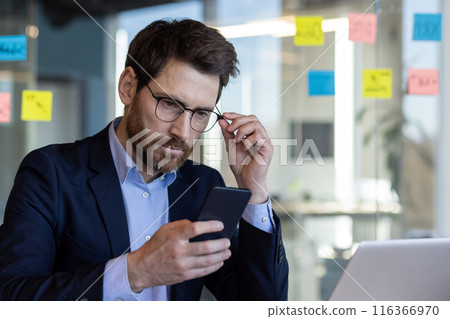 A professional businessman in a suit attentively uses a smartphone, adjusting his glasses in a contemporary office setting with colorful sticky notes in the background. A professional businessman in a suit attentively uses a smartphone, adjusting his glasses in a contemporary office setting with colorful sticky notes in the background. 116366970