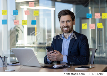 Portrait of successful man in business suit in office interior, mature businessman smiling and looking at camera, satisfied with achievement results, using app on phone. 116366985