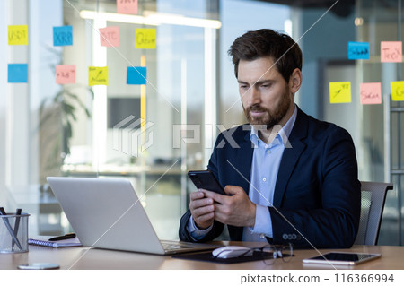 Serious thinking businessman using app on phone, man in business suit mature boss at workplace with laptop inside office, financier investor working. Serious thinking businessman using app on phone, man in business suit mature boss at workplace with laptop inside office, financier investor working. 116366994