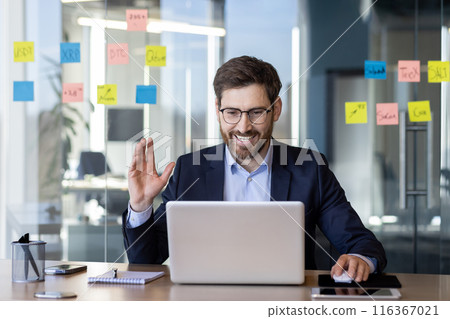 A confident businessman in a suit is smiling and having a video call on his laptop in a modern office. He is seated at a desk with sticky notes on the glass wall behind him A confident businessman in a suit is smiling and having a video call on his laptop in a modern office. He is seated at a desk with sticky notes on the glass wall behind him 116367021