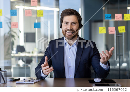 Cheerful businessman wearing headset and talking during a video conference in a modern office. The background features colorful sticky notes on a glass wall. Concept of communication 116367032