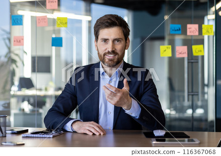 Confident male professional in a business suit gestures while engaging in an animated video call from his sleek, glass-walled office. 116367068