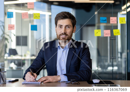 A focused businessman engages in a video call while seated at a desk in a contemporary office setting, surrounded by glass partitions and sticky notes. 116367069