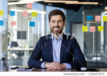 A professional young businessman with a beard and a confident smile, sitting in a well-lit modern office with sticky notes in the background. 116367079