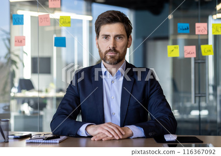 A focused, bearded businessman sits at a glass table in a modern office, surrounded by strategic sticky notes on glass. He projects confidence and professionalism. 116367092