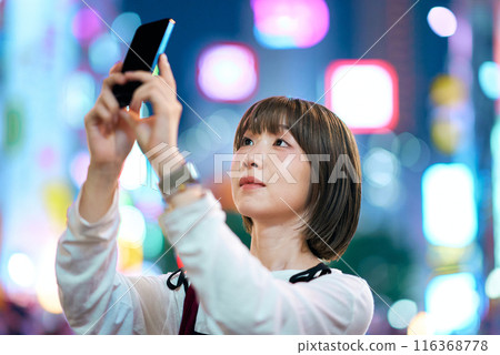 一名年輕女子夜間在市中心舉著智慧型手機 116368778