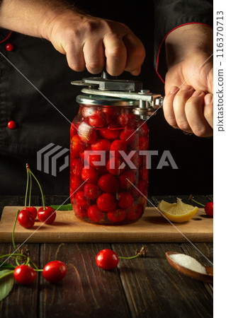 The chef rolls up a jar with a cherry drink or compote with a manual seaming key in his hands. The concept of preserving cherries in jars for future use. Advertising space on the black table The chef rolls up a jar with a cherry drink or compote with a manual seaming key in his hands. The concept of preserving cherries in jars for future use. Advertising space on the black table 116370713
