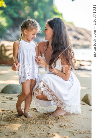 Young woman mother with a little daughter in white dresses on seashore in the shade of trees and palms Young woman mother with a little daughter in white dresses on seashore in the shade of trees and palms 116371031