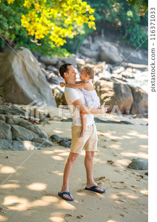 Little girl on father's hands smile and rejoice on seashore in the shade of trees and palms. Happy family concept Little girl on father's hands smile and rejoice on seashore in the shade of trees and palms. Happy family concept 116371033