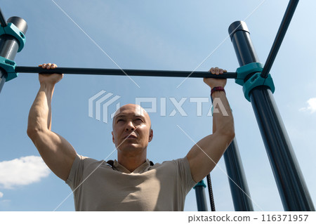 Handsome man does pull-ups on horizontal bar at street on sports ground. Outdoors. Summer. Workout. Morning routine. Handsome man does pull-ups on horizontal bar at street on sports ground. Outdoors. Summer. Workout. Morning routine. 116371957