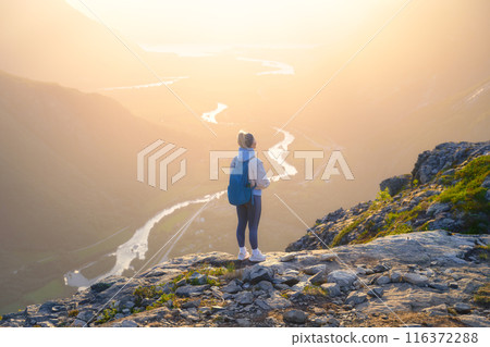 A young athletic girl standing on a rock with a backpack. Sunshine in a mountain valley. 116372288