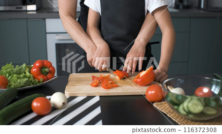 Young mother and her daughter in aprons are meticulously slicing a red bell pepper on a wooden cutting board to prepare a delicious vegetable salad in the kitchen Young mother and her daughter in aprons are meticulously slicing a red bell pepper on a wooden cutting board to prepare a delicious vegetable salad in the kitchen 116372308