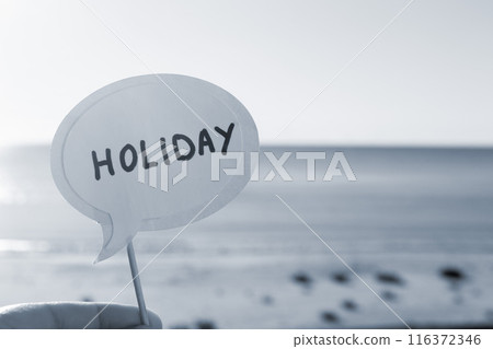 Stick with paper speech bubble with words Holiday on background blue sea, sky, sandy beach on sunny summer day. Text-balloons with text from letters. Concept, symbol, sign vacation travel. Blue color Stick with paper speech bubble with words Holiday on background blue sea, sky, sandy beach on sunny summer day. Text-balloons with text from letters. Concept, symbol, sign vacation travel. Blue color 116372346
