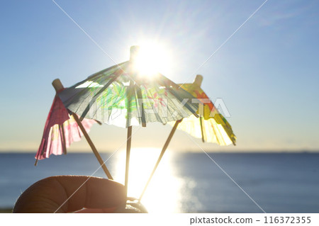 Man holding in hand three paper umbrellas cocktail decorations on background of blue sky, brightly shining sun and sunny path on surface of blue sea. Concept of summer, sea, vacation, travel, tourism Man holding in hand three paper umbrellas cocktail decorations on background of blue sky, brightly shining sun and sunny path on surface of blue sea. Concept of summer, sea, vacation, travel, tourism 116372355