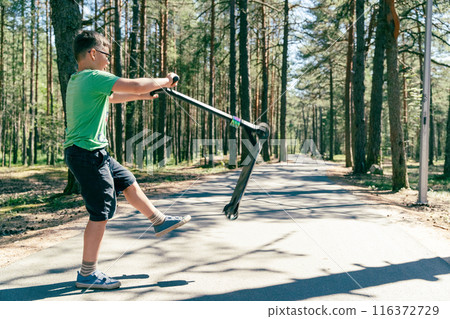 Boy in green shirt and black shorts rides scooter down forest path 116372729