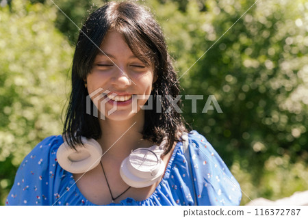 Woman with black hair and blue shirt smiles in park with headphones 116372787