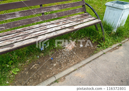 A wooden park bench with visible wear and tear sits next to a trash can A wooden park bench with visible wear and tear sits next to a trash can 116372801